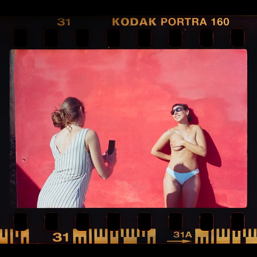 Foto su pellicola: ragazza in slip celeste contro un muro rosso, mentre un'amica la fotografa col telefono