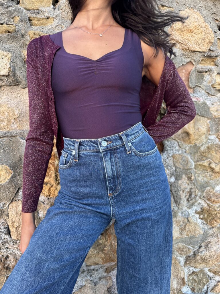 Body viola con cardigan brillante e jeans, davanti a un muro di pietra